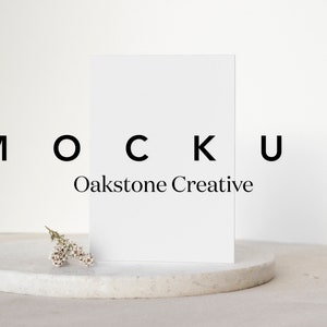 May include: A white card mockup on a white round pedestal with a small sprig of white flowers. The text "MOCKUP Oakstone Creative" is printed below the card.