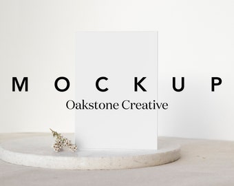 May include: A white card mockup on a white round pedestal with a small sprig of white flowers. The text "MOCKUP Oakstone Creative" is printed below the card.