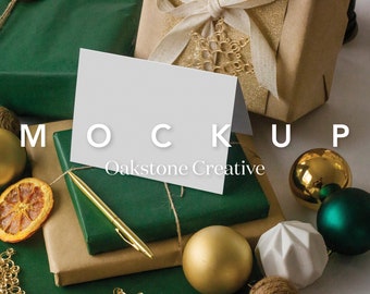 May include: A mockup scene with a blank white card, gold pen, and wrapped gifts. The gifts are wrapped in brown and green paper, tied with twine, and adorned with gold ornaments. The text "MOCKUP" and "Oakstone Creative" are visible.