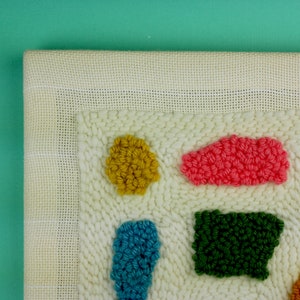 May include: A close-up of a white canvas with colourful yarn pom-poms in various shapes. The pom-poms are arranged in a geometric pattern, creating a modern and abstract design.
