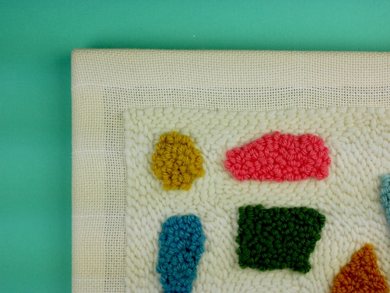 May include: A close-up of a white canvas with colourful yarn pom-poms in various shapes. The pom-poms are arranged in a geometric pattern, creating a modern and abstract design.