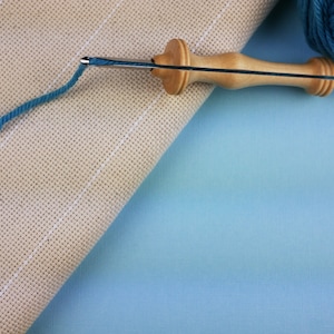 May include: A close-up of a wooden needle threader with a blue yarn tail on a beige Aida cloth. The needle threader is lying on a light blue surface.