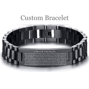 May include: A black metal bracelet with a silver plate engraved with a prayer for pilots. The prayer reads: "Thank you that I have the privilege of flying airplanes. Thank you that I can take to the skies, see earth become small and fall away beneath me. O Lord, help me to fly with skill and diligence, to follow procedure and to keep those I carry safe. Alert me to difficulties and dangers, so that I have time to respond and pray. And as I take to the skies, come lift my heart too. May I always give thanks for the goodness in my life. As the earth blurs away beneath me, come give me your heart for it. Please inspire my mind to see everything through your eyes. And may my prayers be free, dynamic and effective from this perspectivel Lord, lead me closer to you each time I pilot this plane."