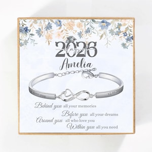 May include: A silver-toned bracelet with an infinity symbol and the name "Amelia" is displayed in a gift box. The box features floral designs and the text "Class of 2026". The bracelet has the inscription "I love you" and "12.10.2024".