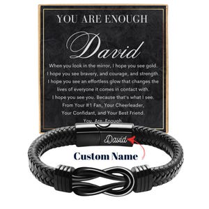 May include: A black braided leather bracelet with a silver-colored infinity knot clasp. The bracelet has a magnetic clasp with the name "David" engraved. A card in the background reads "YOU ARE ENOUGH" and includes a personalized message.