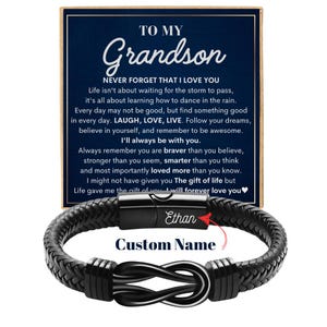 May include: A black braided leather bracelet with a stainless steel infinity symbol clasp. The bracelet is personalized with the name "Ethan" and the words "Custom Name". A navy blue card with the text "To My Grandson" and a loving message is in the background.