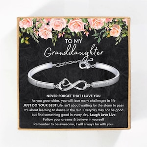 May include: A silver infinity bracelet with a heart charm and the words "I love you" and "12.10.2024" engraved on it. The bracelet is in a box with a floral design and the words "To my Granddaughter" and a message about life and love.