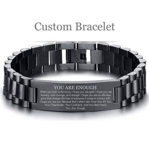 May include: A black stainless steel bracelet with an engraved message that reads "You Are Enough". The message includes a motivational quote about seeing gold, bravery, courage, and strength in yourself. The bracelet is a reminder to be confident and to believe in yourself.