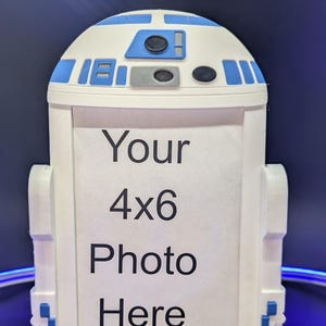 May include: White and blue R2-D2-themed photo frame. The frame is designed to hold a 4x6 photo and features the iconic droid's head and body shape. The text "Your 4x6 Photo Here" is printed on the frame.