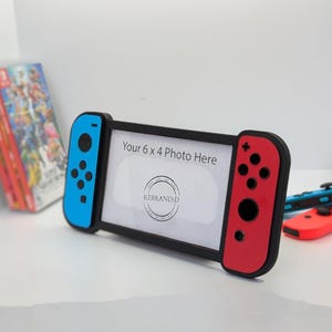 May include: A black Nintendo Switch photo frame with blue and red joy-con controllers. The frame is designed to hold a 6 x 4 inch photo. The text "Your 6 x 4 Photo Here" is printed on the frame.