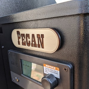 May include: A black smoker with a wooden sign that reads "PECAN" and a digital control panel with a dial and a notice that reads "PRESS AND HOLD DIAL TO SHUTDOWN GRILL".