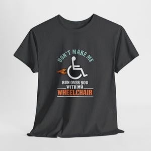 May include: Dark gray t-shirt with the text "DON'T MAKE ME RUN OVER YOU WITH MY WHEELCHAIR". The design includes a wheelchair symbol with flames and text in teal, orange, and white. The shirt is made of a soft material.