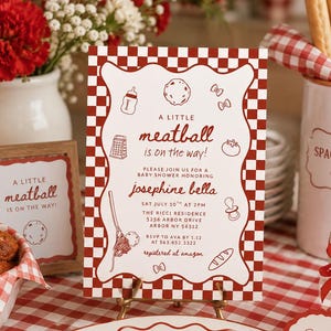 May include: A baby shower table setting with a red and white checkered theme. The centerpiece is a baby shower invitation with the text "A Little Meatball is on the Way!" and a small framed sign with the same text. A bowl of meatballs, flowers, and spaghetti are also visible.