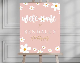 May include: A pink welcome sign with white daisies and gold lettering. The sign says "Welcome to Kendall's 1st birthday party".