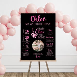 Könnte beinhalten: Schwarzes Geburtstagschild mit rosa Text und einem Foto eines Kindes. Auf dem Schild steht "Chloe, My 2nd Birthday" und enthält Details wie Größe (66 cm), Gewicht (13,6 kg) und Lieblingsdinge. Rosa Ballons umgeben das Schild.