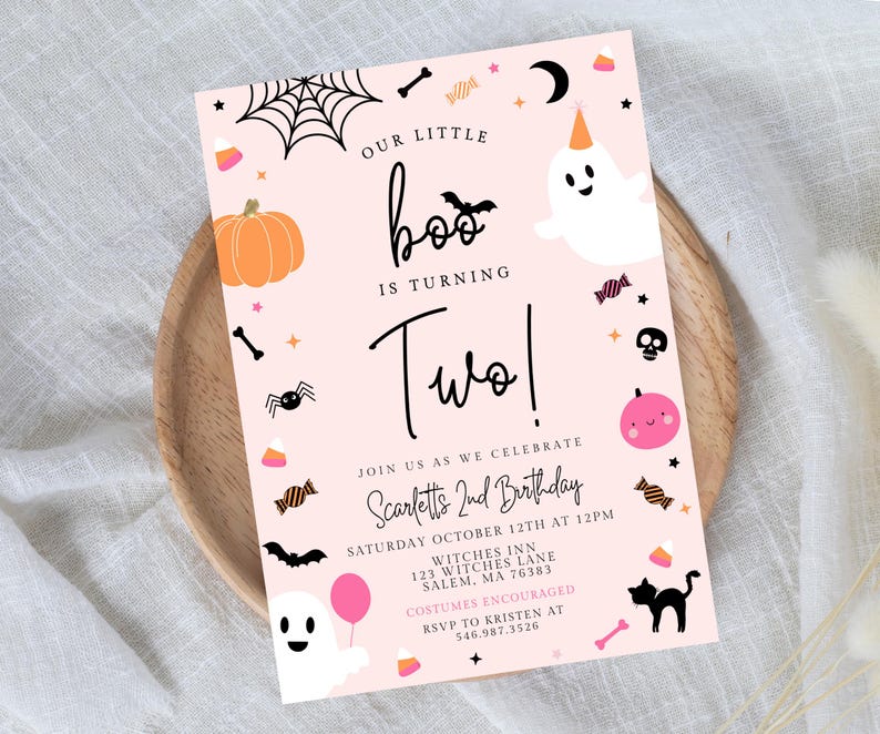 May include: A rectangular birthday invitation card with a light pink background featuring Halloween-themed illustrations including ghosts, a pumpkin, a spider, a bat, a black cat, and candy. The text reads Our Little Boo Is Turning Two!