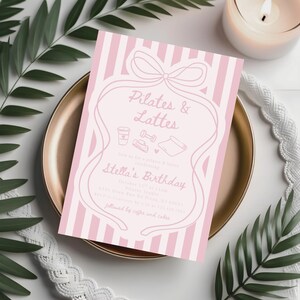 May include: A pink and white striped invitation card with the text "Pilates & Lattes" and "Stella's Birthday". The card features a bow and line art illustrations of a coffee cup, dumbbell, yoga mat, and other fitness-related icons. The invitation is on a gold plate.