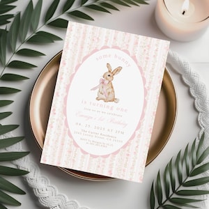 May include: A pink and white invitation card with a bunny illustration. The card reads "some bunny is turning one" and includes event details. The card is on a gold plate with greenery and a candle in the background. The invitation is for a first birthday.