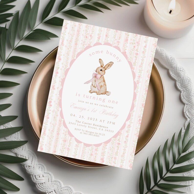 May include: A pink and white invitation card with a bunny illustration. The card reads "some bunny is turning one" and includes event details. The card is on a gold plate with greenery and a candle in the background. The invitation is for a first birthday.