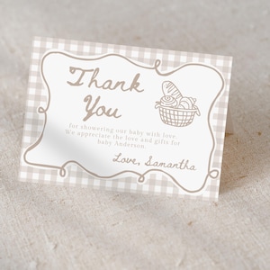 May include: A folded thank you card with a beige gingham pattern background. The card features a decorative border and the words "Thank You" in a handwritten style. A basket of bread is illustrated on the right. The text reads "for showering our baby with love...Love, Samantha."