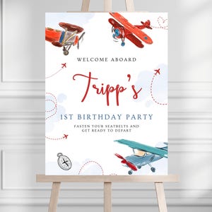 May include: A welcome sign on an easel with a watercolor design featuring red and blue airplanes. The sign reads "Welcome Aboard" and "Tripp's 1st Birthday Party". Decorative dotted lines and airplane graphics surround the text.