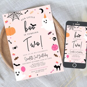 May include: A light pink birthday invitation card and a smartphone displaying the same design. The card features a white ghost, an orange pumpkin, a black spider, a black cat, and text reading Our Little Boo Is Turning Two, Scarlett's 2nd Birthday.