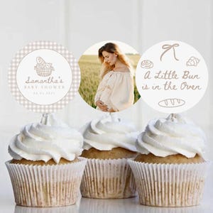 May include: Three cupcakes with white frosting and decorative toppers. One topper has a photo of a pregnant person, another says "A Little Bun is in the Oven", and the last says "Samantha's Baby Shower".