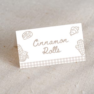 May include: A folded white card with the words "Cinnamon Rolls" in a handwritten font. The card features line drawings of baked goods and a checkered border in shades of beige and brown.