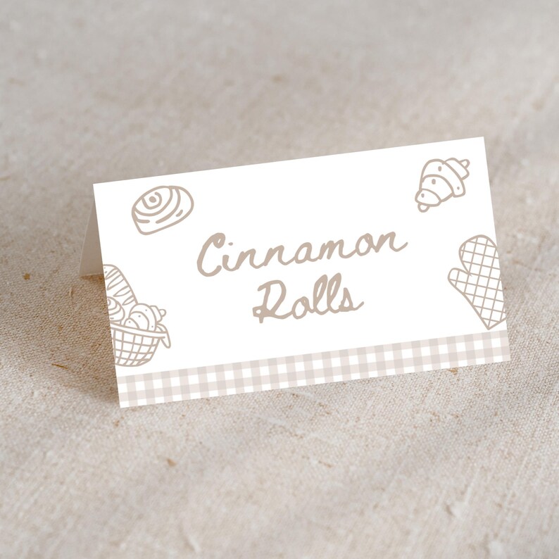 May include: A folded white card with the words "Cinnamon Rolls" in a handwritten font. The card features line drawings of baked goods and a checkered border in shades of beige and brown.