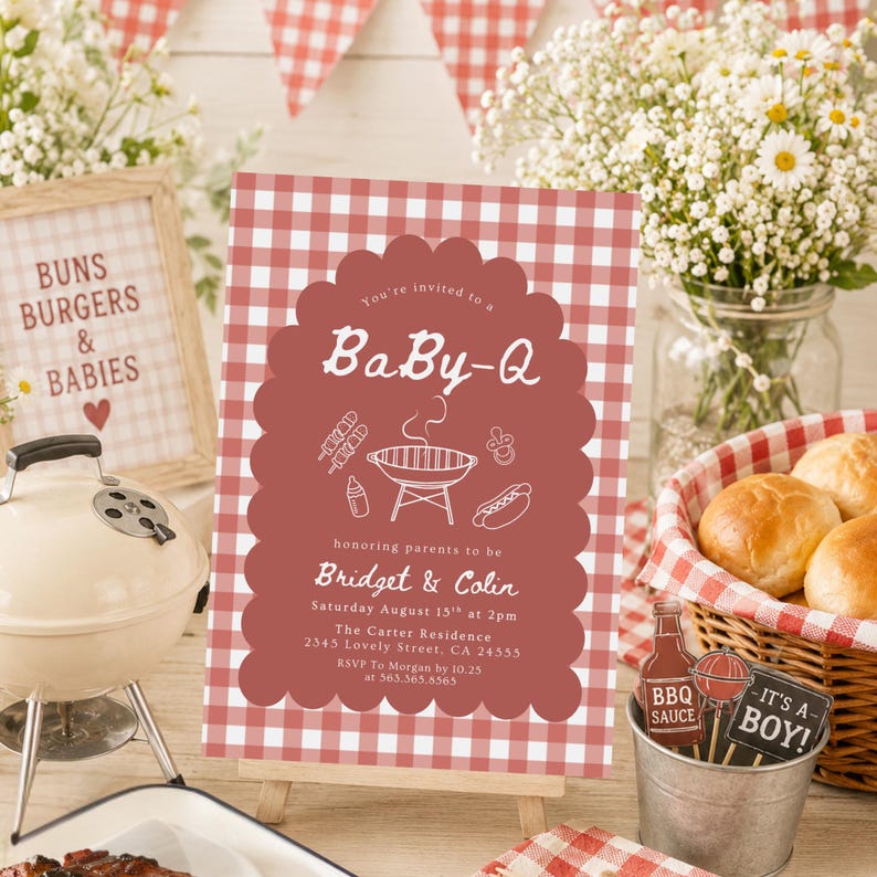 May include: A baby shower invitation with a red and white gingham pattern, featuring the text "BaBy-Q". The invitation is on a small easel, with a miniature grill, flowers, and a basket of rolls in the background. The invitation text reads "Buns, Burgers & Babies".