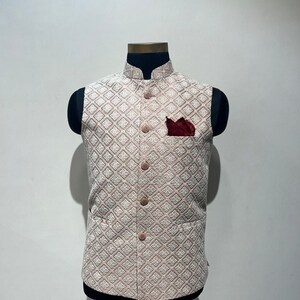 May include: A light pink and white patterned vest with a diamond design, featuring a mandarin collar and matching buttons. A burgundy pocket square is in the chest pocket. The vest is displayed on a black mannequin.