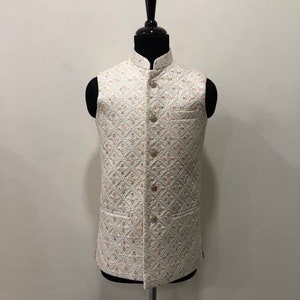 May include: A cream-colored, sleeveless vest with a mandarin collar and a diamond pattern. The vest has a button-down front and two front pockets. The pattern includes gold and pink accents. The vest is displayed on a black mannequin.