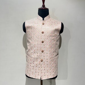 May include: A light pink vest with a mandarin collar and a diamond pattern. The vest has a row of six round, light brown buttons down the front. The fabric has a subtle sheen and a delicate floral design.