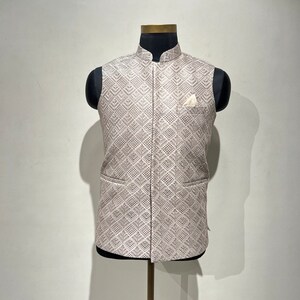 May include: A beige vest with a geometric pattern is displayed on a mannequin. The vest has a mandarin collar, two front pockets, and a pocket square. The pattern is a repeating diamond design in a lighter shade of beige.
