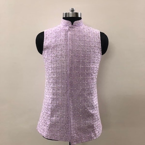 May include: A lavender-colored, sleeveless vest with a mandarin collar. The vest features a grid-like pattern with small, silver-colored embellishments. It is displayed on a black mannequin.