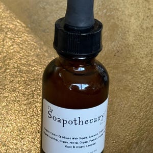 May include: A brown glass bottle with a black dropper and a white label. The label reads "The Soapothecary" and lists organic ingredients. The bottle contains a liquid, likely an oil or serum, for external use.