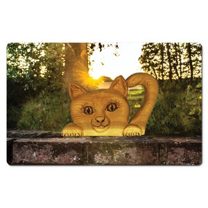 Puede incluir: Escultura de gato de madera marrón claro con una expresión amigable y una cola rizada. El gato está posado en un muro de ladrillo. El fondo presenta una puesta de sol con árboles y vegetación.