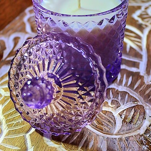 May include: A purple glass candle jar with a lid. The jar has a diamond pattern and the lid has a floral design. The candle is white and has a wick.