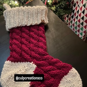 May include: A hand-knitted Christmas stocking featuring a deep red cable knit pattern on the main body, with a thick cream-coloured cuff, heel, and toe. A small brown tag is attached to the cuff, and the text @culpcreationsco is visible.