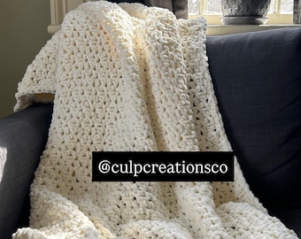 Può includere: Una coperta all'uncinetto color bianco sporco, spessa e con un motivo a punti annodati testurizzati. La coperta presenta un'etichetta rettangolare nera con il testo bianco @culpcreationsco centrato sul tessuto, che mostra una struttura in filato spesso.