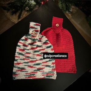 May include: Two crocheted hanging kitchen towels with button closures. One towel features a variegated pattern of red, green, and white yarn, while the other is solid red. The text @culpcreationsco is visible on the variegated towel.