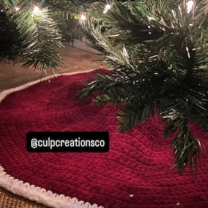 May include: A round, deep red crocheted Christmas tree skirt featuring a thick, textured white border trim. The fabric has a soft, plush appearance. A black rectangular label with the white text @culpcreationsco is attached to the surface.