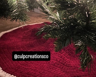 Puede incluir: Una falda de árbol de Navidad redonda de ganchillo en color rojo oscuro con un borde blanco grueso y texturizado. La tela tiene un aspecto suave y afelpado. Una etiqueta rectangular negra con el texto blanco @culpcreationsco está fijada.