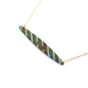 Puede incluir: Un collar de cadena de oro con un colgante turquesa y dorado. El colgante es un rect&aacute;ngulo largo y delgado con un patr&oacute;n de espiral.