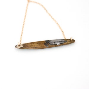 Puede incluir: Un collar de cadena dorada con un colgante largo y delgado de lat&oacute;n. El colgante tiene una forma ligeramente curvada y una superficie rugosa y texturizada.