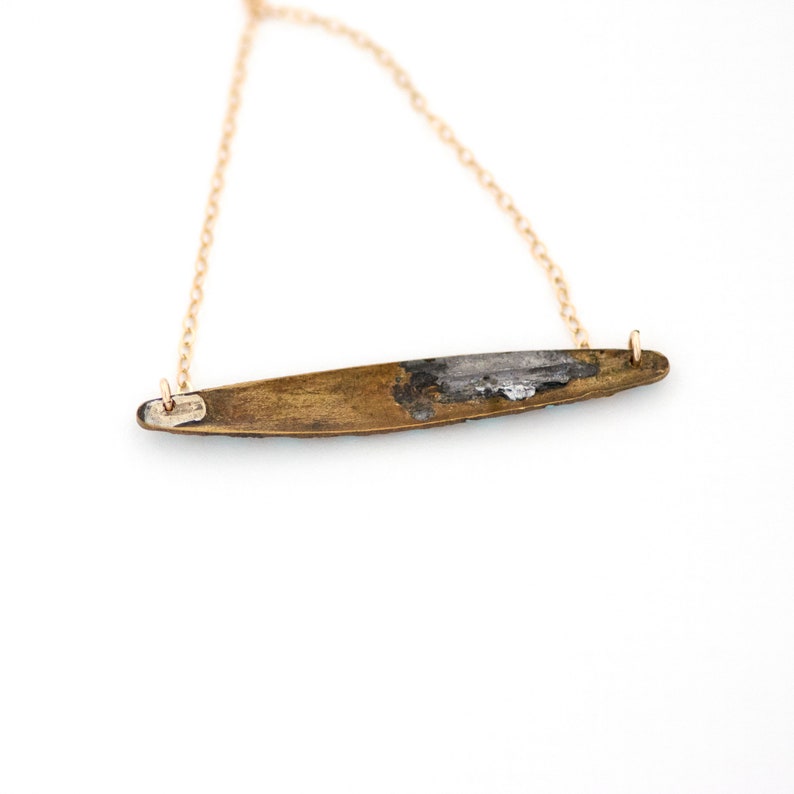 Puede incluir: Un collar de cadena dorada con un colgante largo y delgado de lat&oacute;n. El colgante tiene una forma ligeramente curvada y una superficie rugosa y texturizada.
