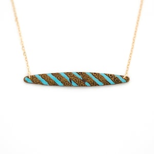 Puede incluir: Un collar de cadena dorada con un colgante de barra turquesa y dorada. La barra tiene un patr&oacute;n de rayas y un dise&ntilde;o intrincado.