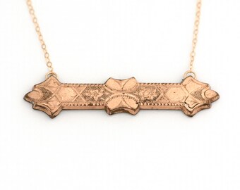 Puede incluir: Un collar de oro con una cadena delicada y un colgante rectangular largo. El colgante está grabado con un patrón floral.