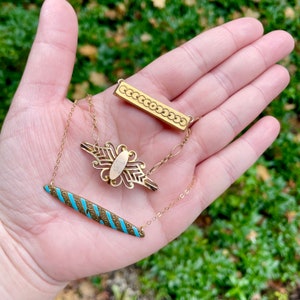 Puede incluir: Un collar de oro con tres colgantes. Los colgantes son una barra rectangular con un dise&ntilde;o de c&iacute;rculo repetitivo, un colgante con forma de mariposa con un &oacute;valo dorado en el centro y una barra larga y delgada con un dise&ntilde;o de espiral azul y dorado.