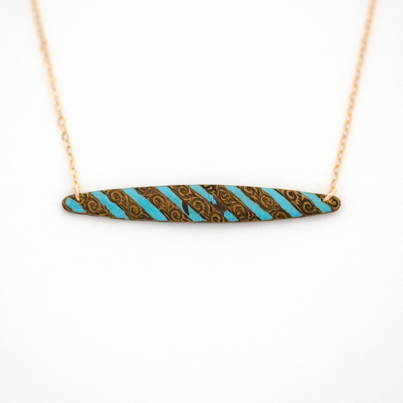 Puede incluir: Un collar de cadena dorada con un colgante de rayas turquesa y marr&oacute;n. El colgante es rectangular y tiene una superficie texturizada.